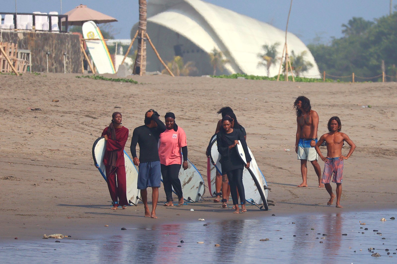 Students in modest swimwear carrying boards back up the beach with their instructors