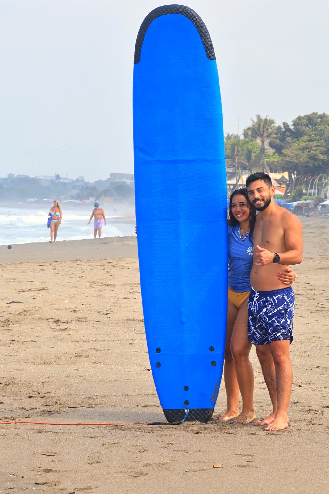 Smiling couple standing with a tall blue surfboard after their lesson
