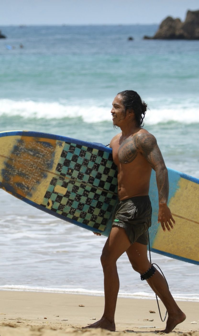 Gov walking up the beach carrying his surfboard on a surf trip