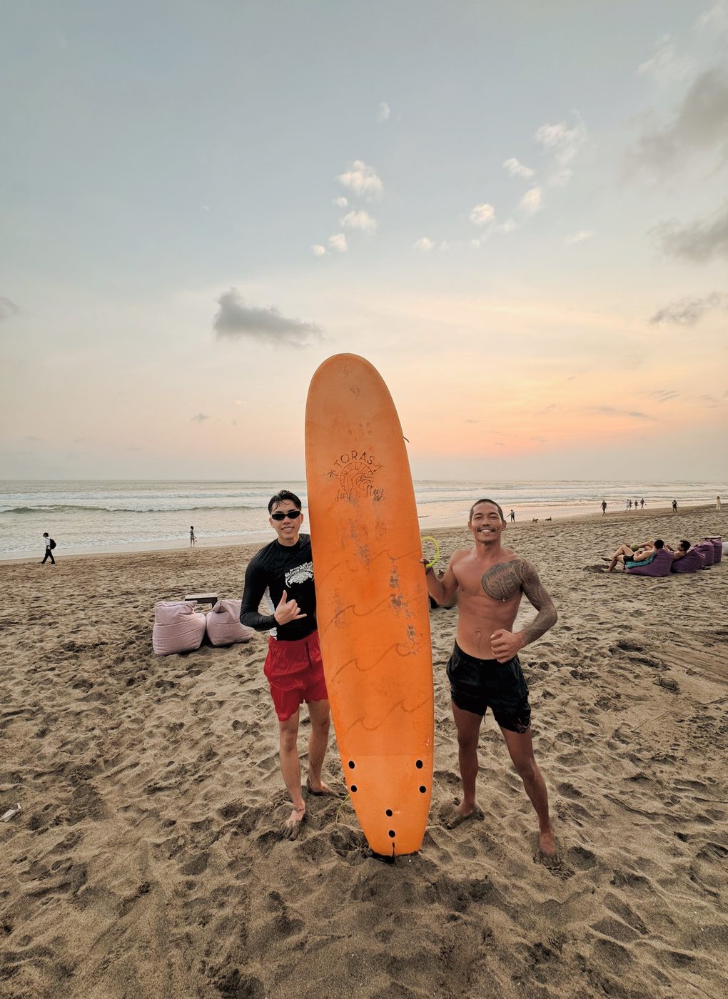 Gov and a student posing with a surfboard on the beach at sunset