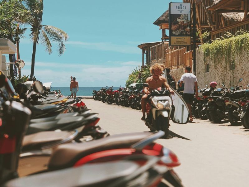 A beach street in Bali lined with scooters, a surfer riding past with a board